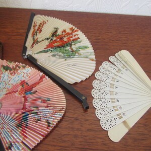 May include: Three vintage folding fans. One fan is white with a black lace design, one is pink with a floral design, and one is white with a floral design.