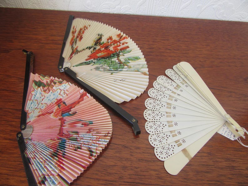 May include: Three vintage folding fans. One fan is white with a black lace design, one is pink with a floral design, and one is white with a floral design.