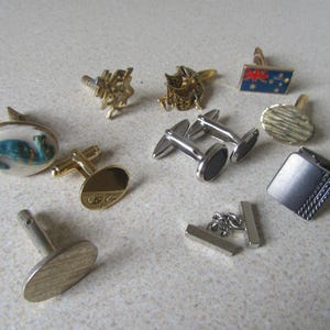 May include: A collection of various cufflinks in gold and silver tones. Designs include oval, rectangular, and novelty shapes. Some cufflinks feature patterns, while others have enamel details in blue, white, and red.