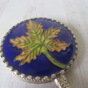 Peut inclure: Miroir de poche rond avec un motif de feuille sur fond bleu. La feuille est verte et orange, avec un contour argenté. Le miroir a une poignée argentée.