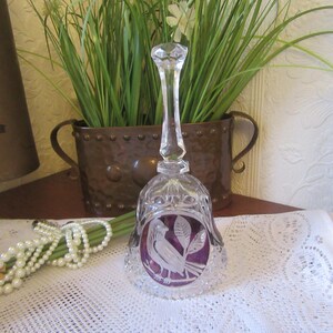 May include: A clear glass bell with a long stem and a faceted top. The bell has a purple insert with a bird and leaf design. The bell is on a white lace tablecloth.