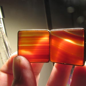 Puede incluir: Un par de gemelos cuadrados con una paleta de colores cálidos de naranja, rojo y amarillo. Los gemelos tienen un patrón rayado y arremolinado, encerrados en un marco metálico delgado. El diseño recuerda a la piedra natural.