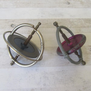 May include: Two antique gyroscopes. One has a silver metal frame with a dark gray disc and a brown spindle. The other has a gray frame with a pink disc and a brown spindle. Both are on a light wood surface.