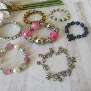 May include: Collection of beaded bracelets in various styles and colors. Includes bracelets with pink beads, silver accents, and star charms. Other bracelets feature gold, clear, and dark blue beads. A cross charm is on one bracelet.