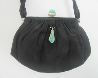 Könnte beinhalten: Eine schwarze Stoffhandtasche mit gerafftem Design und einem Tragegriff. Der Verschluss ist mit einem abgerundeten grünen Stein und einer Reihe kleiner klarer Kristalle verziert. Ein dekorativer silberfarbener Metallanhänger mit grünem Fächer.