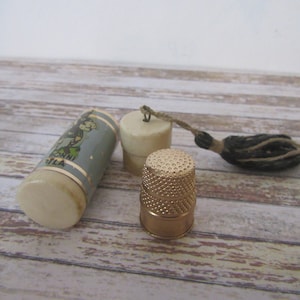 May include: A vintage sewing kit with a white and blue cylindrical case with a cartoon image and text that reads "RONNI FILA", a white thimble with a gold top, and a black and brown tassel.