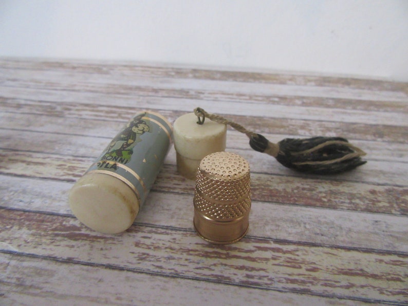 May include: A vintage sewing kit with a white and blue cylindrical case with a cartoon image and text that reads "RONNI FILA", a white thimble with a gold top, and a black and brown tassel.