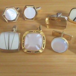 May include: A collection of vintage cufflinks in various shapes and designs. Gold-toned metal cufflinks with mother-of-pearl, geometric, and rectangular designs are displayed on a wooden surface. Some have a single gemstone.