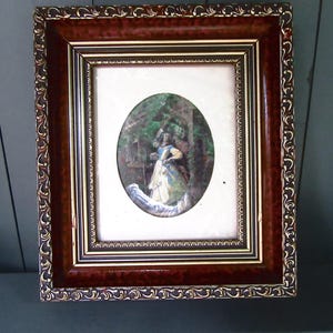 Op de afbeelding: Een antiek, omlijst ovaal portret met een vrouw in een witte jurk met blauwe accenten, tegen een bosachtergrond. De lijst is een rijk, donkerbruin met gouden details en sierlijke gravures, wat een vintage esthetiek creëert.