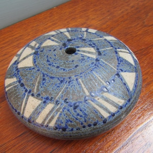 Könnte beinhalten: Eine runde Keramikvase mit blauem und weißem geometrischen Muster. Die Vase hat ein zentrales Loch und steht auf einer Holzoberfläche. Das Design zeigt Spiral- und Dreiecksformen.