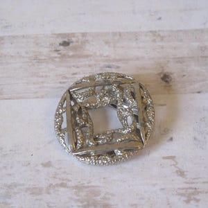 K&ouml;nnte beinhalten: Eine silberfarbene Brosche aus Metall mit quadratischem Design und filigranen, miteinander verwobenen Details. Die Brosche hat ein zartes, durchbrochenes Muster.