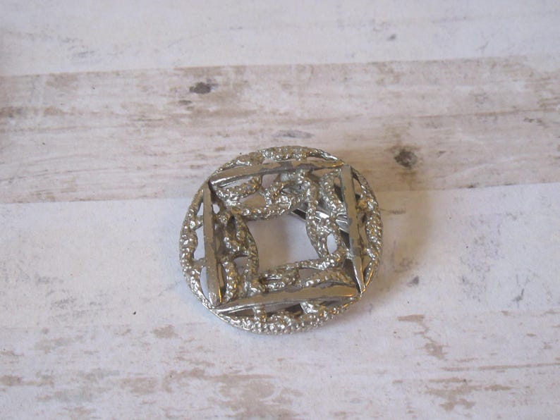 K&ouml;nnte beinhalten: Eine silberfarbene Brosche aus Metall mit quadratischem Design und filigranen, miteinander verwobenen Details. Die Brosche hat ein zartes, durchbrochenes Muster.