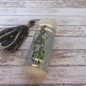 May include: A white and blue cylindrical bottle stopper with a gold trim and a label featuring a cartoon man in a kilt. The label also reads "Bonni O'Tla". The bottle stopper has a black and white tassel.