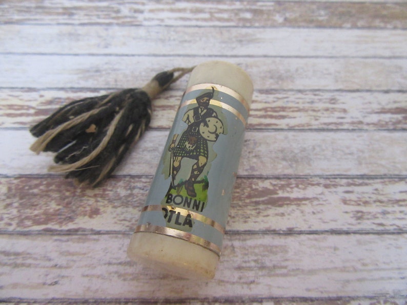 May include: A white and blue cylindrical bottle stopper with a gold trim and a label featuring a cartoon man in a kilt. The label also reads "Bonni O'Tla". The bottle stopper has a black and white tassel.