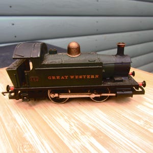 May include: A dark green and black model train with the words "GREAT WESTERN" in red lettering. The model features a brass dome and a black smokestack. The train is displayed on a wooden surface.