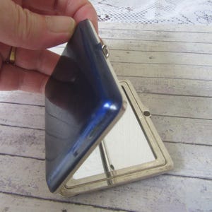 May include: A vintage, blue compact mirror with a silver-toned frame. The compact is open, revealing a mirror. The compact is held by a hand, showing the shiny, reflective surface. The compact is on a light-colored wooden surface.