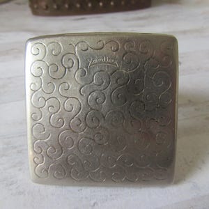 Op de afbeelding: Een vierkante, zilverkleurige metalen doos met afgeronde randen. Het oppervlak is bedekt met een reliëf, wervelend patroon. Het woord "Yardley" is cursief ingeschreven. Waarschijnlijk een vintage item.