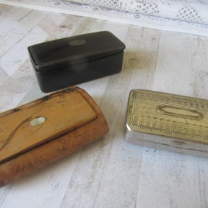May include: Three antique boxes of varying styles. A black rectangular box with a small oval accent, a light brown wooden box with a hinged lid and a silver rectangular box with a textured surface and an oval accent.