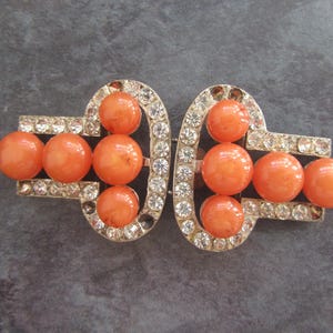 May include: Vintage Art Deco style brooch with a silver-tone metal setting. The brooch features orange, round beads and is encrusted with clear rhinestones. The brooch is a two-piece design.