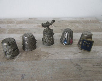 May include: Five silver thimbles with different designs. One thimble features a genie lamp, another has a crown, one has a blue and red jewel, one has a Holy Bible design, and the last one has a building design.