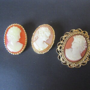 May include: Three cameo brooches with gold-tone settings. The cameos feature a white profile of a woman on a peach-colored background.