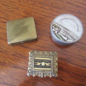 Op de afbeelding: Drie kleine, decoratieve doosjes. Eén is een gouden metalen doosje met een scharnierend deksel. Een ander is een rond, zilveren doosje met een zwart-wit afbeelding van een stad en de tekst "SOUVENIR OF CLIFTONVILLE". De derde is een gouden metalen doosje met een decoratief, versierd ontwerp.