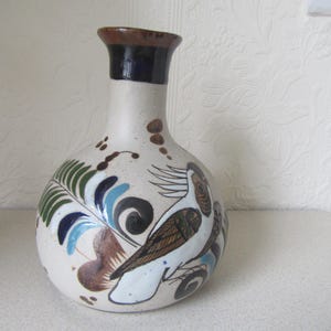 May include: A white ceramic vase with a brown, blue and green painted design of a bird with a long tail. The vase has a narrow neck and a wide base.