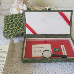 May include: An open, rectangular, green and gold box with a red interior, containing calligraphy and a magnifying glass. A smaller, matching box is visible to the left. A page of text is in the foreground.