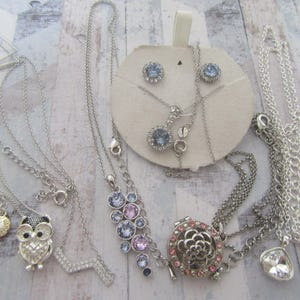 May include: A set of silver necklaces with various charms, including an owl, a flower, and a crystal cluster. The necklaces are displayed on a white background with a card showing a pair of silver earrings with blue crystals.