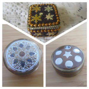 May include: A collection of three small decorative boxes. One is a square box covered in dark beads, gold trim, and gold snowflake accents. The other two are round metal boxes, one with a white and gold mandala pattern and one with a star and circles.