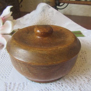 Peut inclure: Un petit bol rond en bois avec un couvercle. Le bol et le couvercle sont de couleur marron chaud. Le couvercle a un petit bouton arrondi. Le bol est posé sur un tissu en dentelle blanche. Une fleur rose et blanche est en arrière-plan.