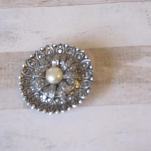 K&ouml;nnte beinhalten: Eine silberne Brosche mit einer Perle in der Mitte und einem detaillierten Blumendesign.