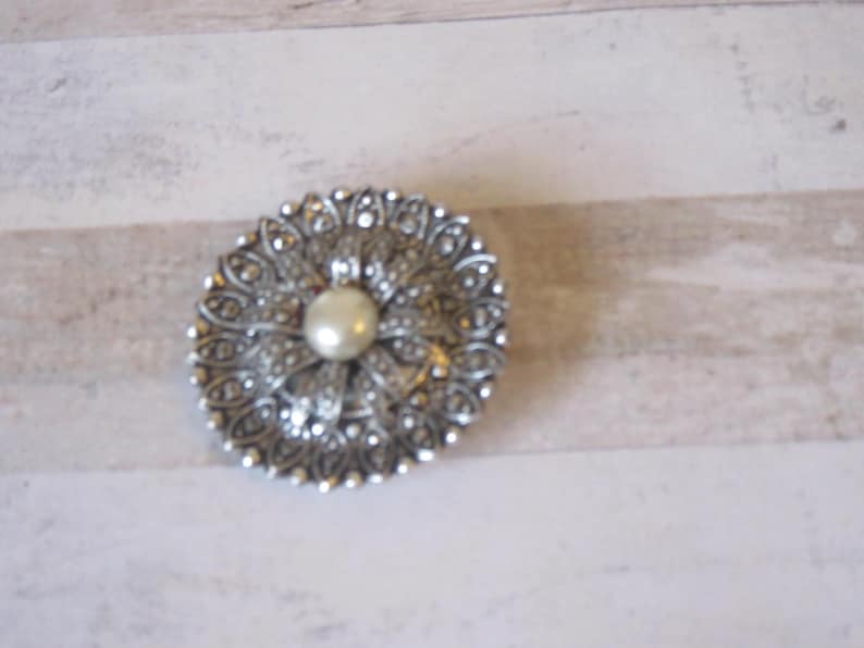 K&ouml;nnte beinhalten: Eine silberne Brosche mit einer Perle in der Mitte und einem detaillierten Blumendesign.