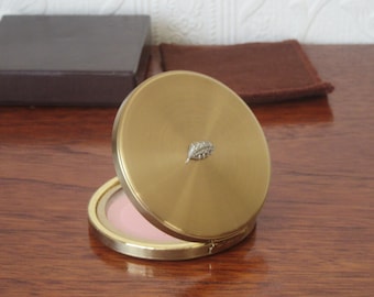 May include: A gold compact with a leaf design on the lid. The compact is open and shows a pink powder inside.