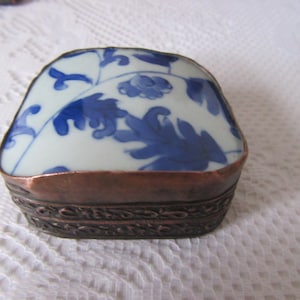Op de afbeelding: Een klein vierkant doosje met een blauw en wit porseleinen deksel en een koperen basis. Het deksel heeft een bloemenmotief.