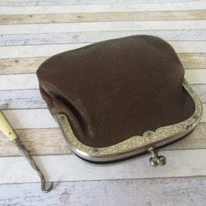 May include: A brown leather coin purse with a decorative silver clasp and a small metal hook tool with a cream-colored handle. The purse has a textured surface and a curved shape.