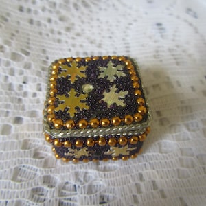 May include: A small, square decorative box with a dark purple beaded exterior. Gold-colored snowflake designs and small gold beads adorn the box. The edges are trimmed with silver and gold beads. A small, clear gem is centered on the top.