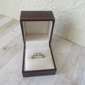 May include: A gold ring with three clear stones, displayed in a brown jewelry box with a white interior. The box is open, revealing the ring against a soft, white fabric. The ring is centered in the box.