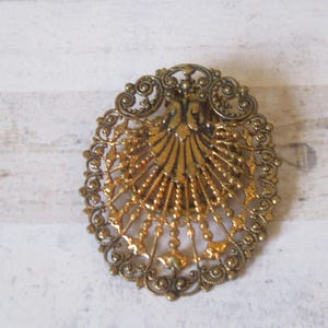 K&ouml;nnte beinhalten: Eine goldfarbene Filigranbrosche mit einem detaillierten Design, das eine f&auml;cherf&ouml;rmige Mitte zeigt.