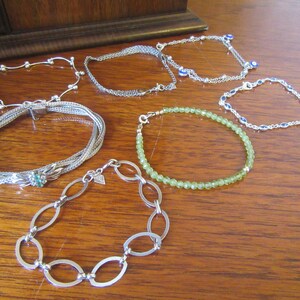 Può includere: Una collezione di braccialetti d'argento con vari design, tra cui un braccialetto a catena, un braccialetto con perline, un braccialetto con una pietra verde e un braccialetto con una catena delicata.