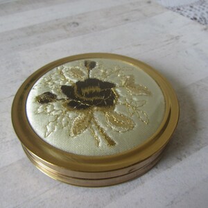 Könnte beinhalten: Ein runder, vintage Kompaktpuder mit einem goldfarbenen Metallgehäuse. Der Deckel zeigt ein gesticktes Blumendesign mit einer dunkelbraunen Rose und goldfarbenen Blättern auf cremefarbenem Hintergrund. Der Puder ist geschlossen und liegt auf einer hellen Holzoberfläche.