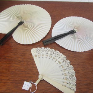 May include: Three white folding fans. Two fans are made of paper with black handles. The third fan is made of white plastic with a delicate lace design and a white handle.