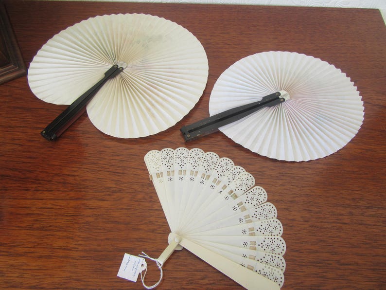 May include: Three white folding fans. Two fans are made of paper with black handles. The third fan is made of white plastic with a delicate lace design and a white handle.