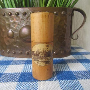 Puede incluir: Un recipiente cilíndrico de madera con una imagen de paisaje en la parte delantera. El recipiente es de color marrón claro y se encuentra sobre una superficie a cuadros azules y blancos. En el fondo hay una olla de color cobre con asas.