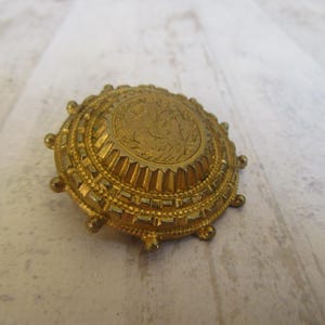 Antik brosch från ca 1920-talet. Guldfärgad rund brosch med etsad blomma. Okänt tillverkarstämpel.