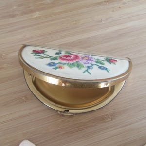 May include: A vintage, half-circle compact with a gold metal frame and a white embroidered floral design. The compact is open, revealing a gold-colored interior.