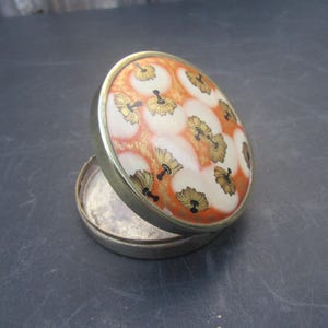 Könnte beinhalten: Ein runder Vintage-Kompaktpuder mit floralem Design. Der Deckel zeigt ein Muster aus orangefarbenen, goldenen und schwarzen Blumenmotiven auf weißem Hintergrund. Der Kompaktpuder hat einen silberfarbenen Metallrahmen und eine Scharnieröffnung.