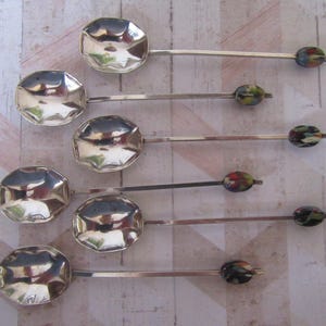 Könnte beinhalten: Ein Set aus sechs silberfarbenen Metalllöffeln mit facettierten, achteckigen Laffen und langen, schlanken Stielen. Jeder Stiel endet in einer dekorativen, ovalen Perle mit einem mehrfarbigen, gesprenkelten Muster in Grün, Rot und Schwarz.