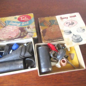 May include: Vintage Tala Icing Set with boxes and tools. The boxes have illustrations of decorated cakes. The set includes metal icing tips, a metal cylinder, and other tools. The boxes are yellow and brown.