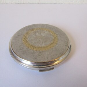 May include: A silver compact with a gold-colored wreath design on the lid.